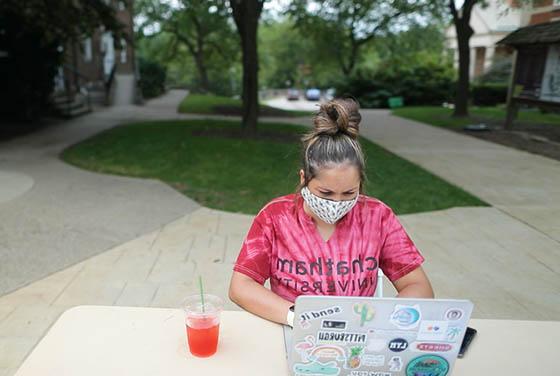 Photo of a masked female student in a Chatham University shirt, working on her laptop outside on Shadyside Campus